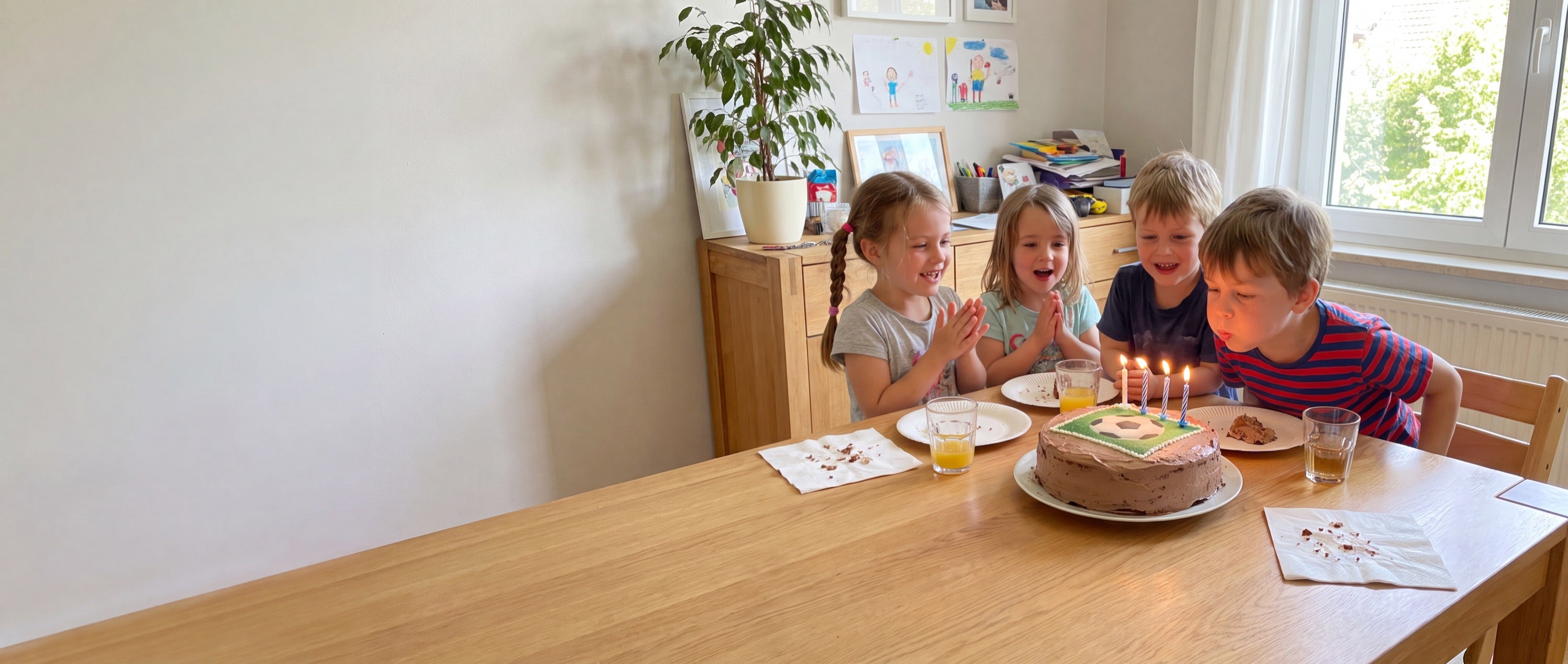 Vier Kinder an einem Geburtstagstisch, ein Junge pustet die Kerzen auf einer Torte mit essbarem Fußball-Foto aus
